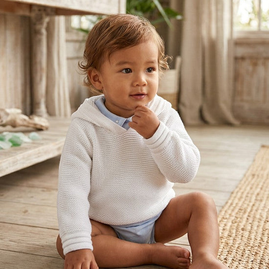 Carriage Boutique baby boy wearing a soft white knit hoodie with an attached hood, sitting on a wooden floor.