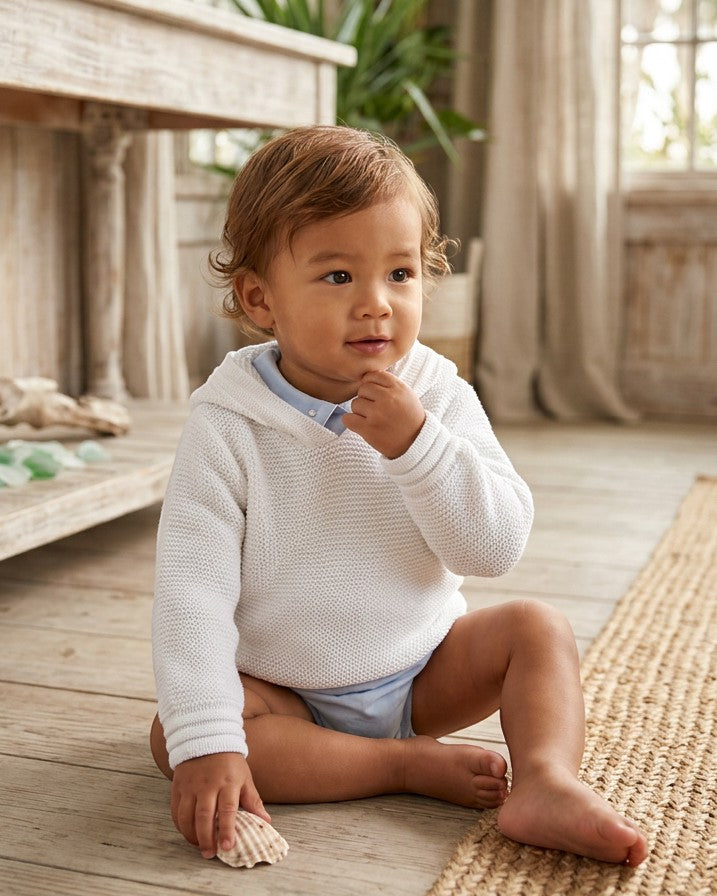 Carriage Boutique baby boy wearing a soft white knit hoodie with an attached hood, sitting on a wooden floor.