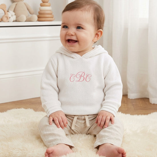 Carriage Boutique white knit baby hoodie with pink monogram embroidery, worn by a smiling baby on a sheepskin rug.