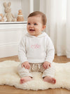 Carriage Boutique white knit baby hoodie with pink monogram embroidery, worn by a smiling baby on a sheepskin rug.