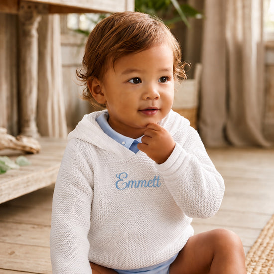 Carriage Boutique white knit baby hoodie with blue embroidered name on chest worn by a toddler in a rustic room