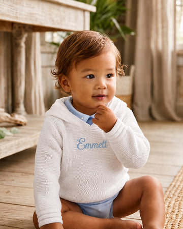 Carriage Boutique white knit baby hoodie with blue embroidered name on chest worn by a toddler in a rustic room