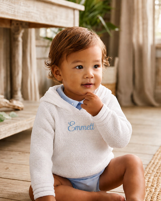 Carriage Boutique white knit baby hoodie with blue embroidered name on chest worn by a toddler in a rustic room