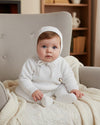 Carriage Boutique baby boy wearing a white knit baptism outfit with a matching bonnet, sitting in a cozy armchair.