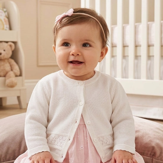 Carriage Boutique baby girl wearing a classic white knit cardigan with button closure over a soft pink dress