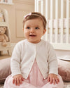 Carriage Boutique baby girl wearing a classic white knit cardigan with button closure over a soft pink dress
