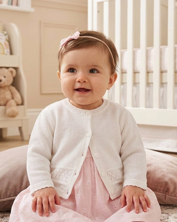 Carriage Boutique baby girl wearing a classic white knit cardigan with button closure over a soft pink dress