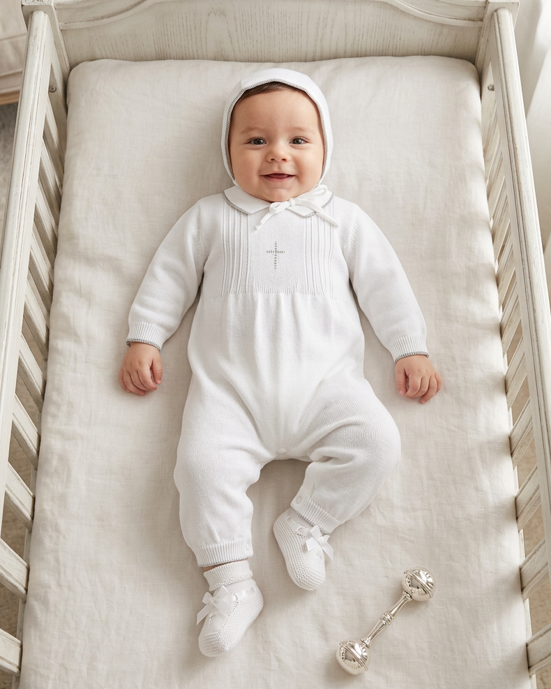 Baby boy wearing a Carriage Boutique white knit christening romper with silver cross embroidery and matching bonnet.