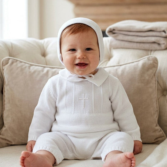 Baby boy wearing a white Carriage Boutique knit cotton christening outfit with a matching bonnet and embroidered cross.