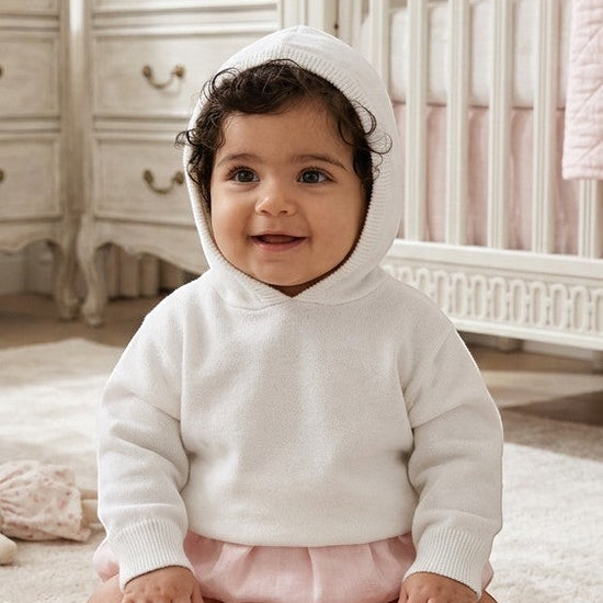 Carriage Boutique white knit hooded baby sweater made of soft cotton worn by a smiling baby in a nursery.