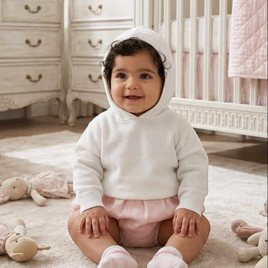 Carriage Boutique white cotton knit hooded baby sweater worn by a smiling baby in a nursery