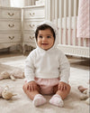 Carriage Boutique white cotton knit hooded baby sweater worn by a smiling baby in a nursery