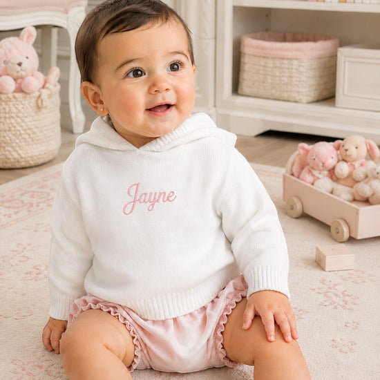 Carriage Boutique white knit hooded baby sweater with personalized pink embroidery, worn by a smiling baby girl.