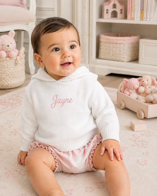 Carriage Boutique white knit hooded baby sweater with personalized pink embroidery, worn by a smiling baby girl.