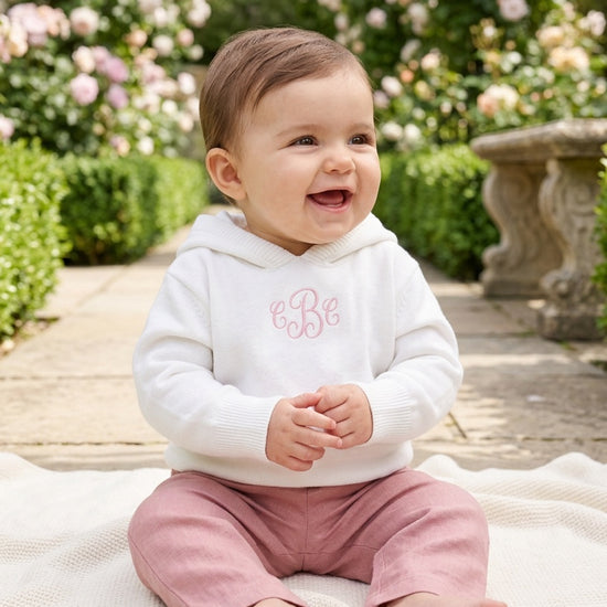 Carriage Boutique white hooded knit baby sweater with pink monogram, worn by a smiling baby in a garden setting.