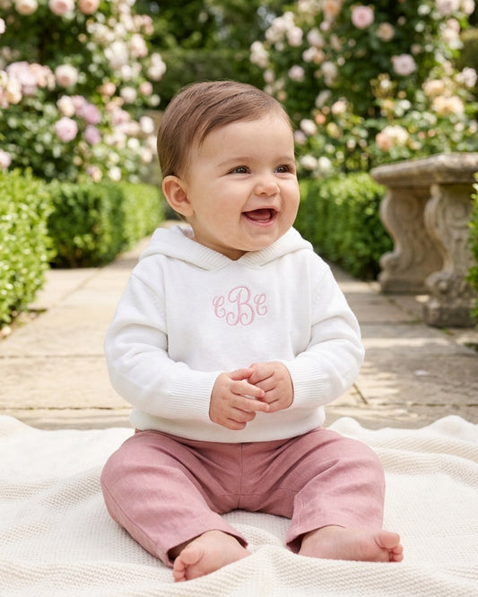 Carriage Boutique white hooded knit baby sweater with pink monogram, worn by a smiling baby in a garden setting.