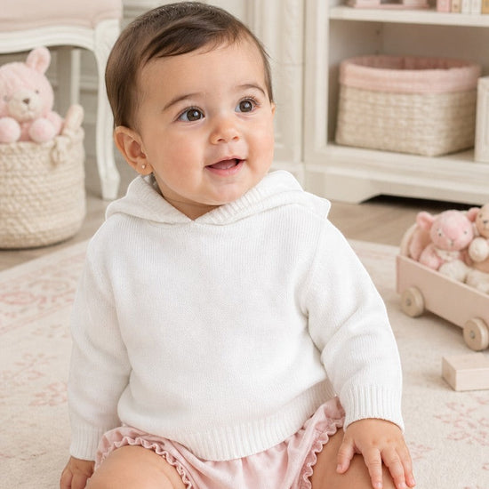Carriage Boutique white knit hooded sweater worn by a smiling baby girl sitting on a soft rug in a nursery.