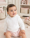 Carriage Boutique white knit hooded sweater worn by a smiling baby girl sitting on a soft rug in a nursery.