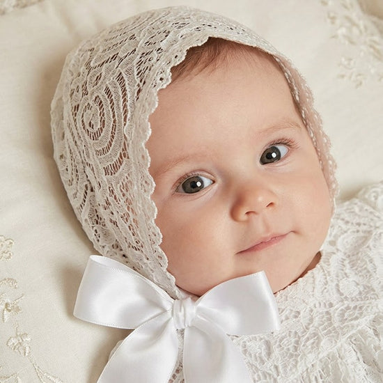 Carriage Boutique baby girl wearing an elegant white lace baptism bonnet with a soft satin ribbon tie.