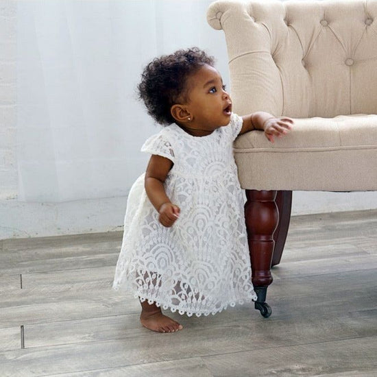 Carriage Boutique white lace baptism dress with matching bonnet worn by a toddler standing by a tufted chair