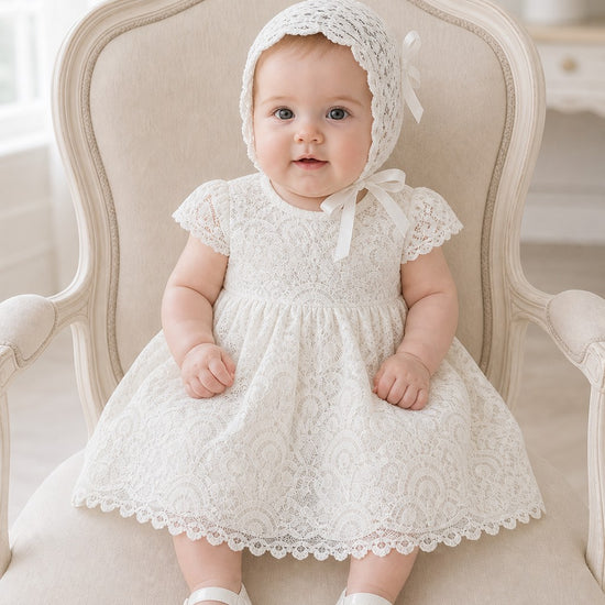 Baby girl wearing a white Carriage Boutique lace baptism dress and matching bonnet while sitting in a vintage chair.