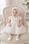 Baby girl wearing a white Carriage Boutique lace baptism dress and matching bonnet while sitting in a vintage chair.