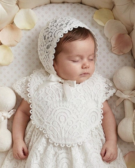 Carriage Boutique white lace christening bib worn by a sleeping baby in a wicker bassinet with a matching bonnet