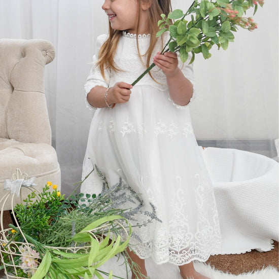 Carriage Boutique white long sleeve lace christening gown with scalloped hem worn by a toddler holding flowers