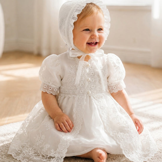 Carriage Boutique baby girl in a white lace christening gown and matching bonnet sitting on a carpeted floor.