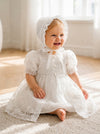 Carriage Boutique baby girl in a white lace christening gown and matching bonnet sitting on a carpeted floor.