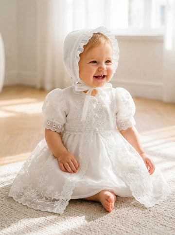 Carriage Boutique baby girl in a white lace christening gown and matching bonnet sitting on a carpeted floor.