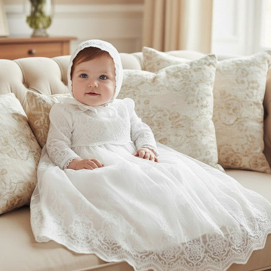 Baby girl wearing a Carriage Boutique white long sleeve lace christening gown with matching bonnet on a sofa