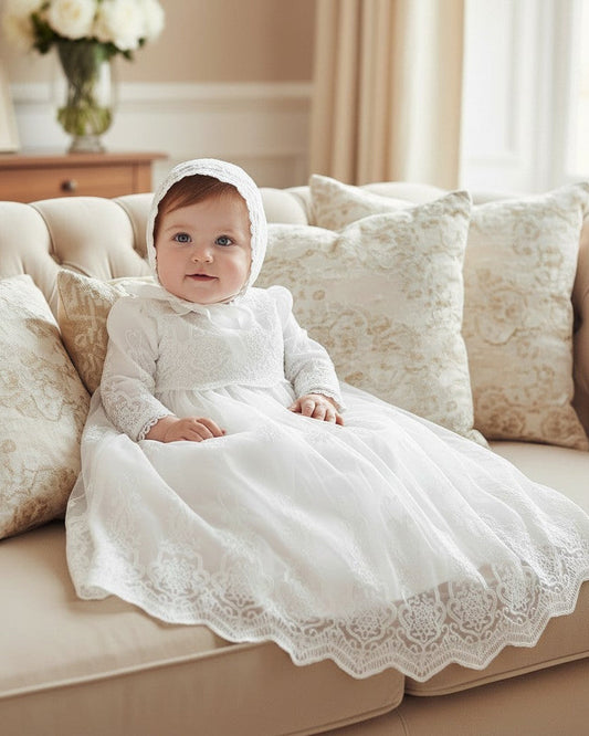 Baby girl wearing a Carriage Boutique white long sleeve lace christening gown with matching bonnet on a sofa
