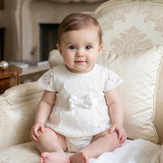 Carriage Boutique baby girl white lace christening bubble romper with a satin waist ribbon sitting in an armchair