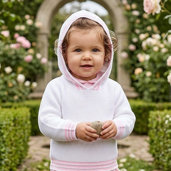 Carriage Boutique baby girl wearing a white hooded sweater with pink striped trim in a lush garden setting.