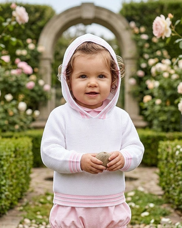 Carriage Boutique baby girl wearing a white hooded sweater with pink striped trim in a lush garden setting.