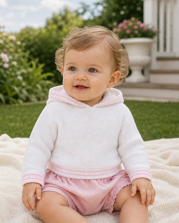 Carriage Boutique baby girl wearing a white hooded sweater with pink trim and matching pink shorts outdoors.