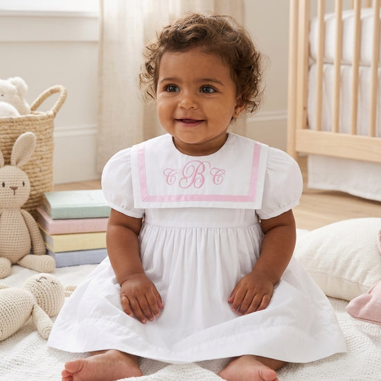Toddler wearing a Carriage Boutique white and pink short sleeve dress with a monogrammed sailor collar in a nursery.