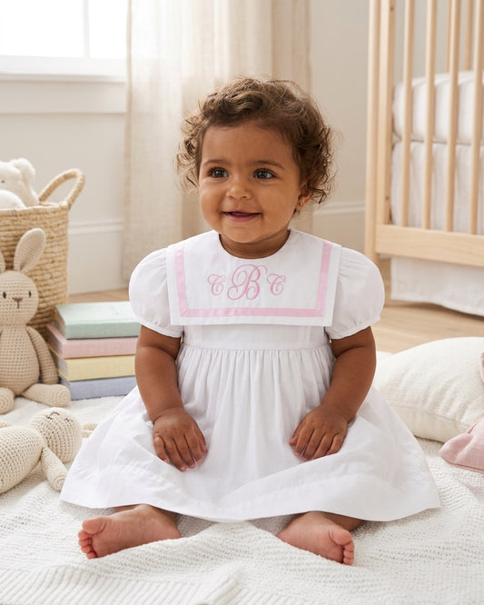 Toddler wearing a Carriage Boutique white and pink short sleeve dress with a monogrammed sailor collar in a nursery.