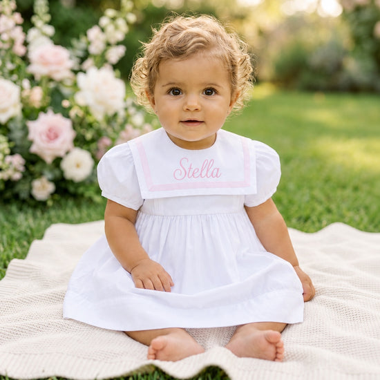 Carriage Boutique white and pink short sleeve toddler dress with personalized embroidery sitting on a garden blanket