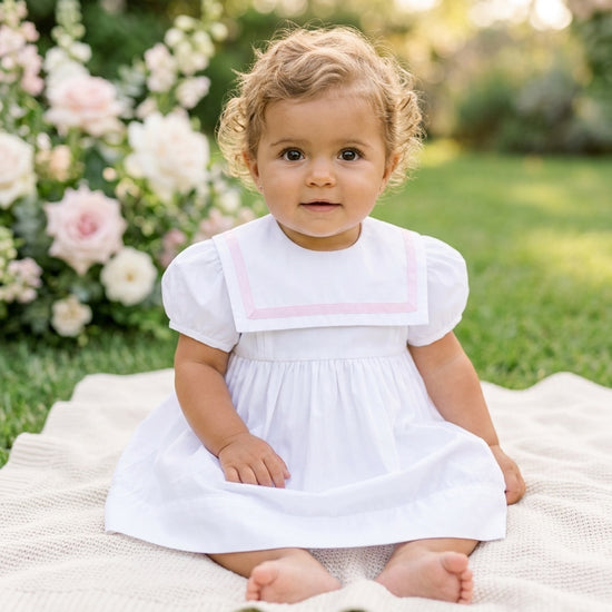 Toddler wearing a Carriage Boutique white and pink short sleeve sailor-style dress sitting on a blanket in a garden.