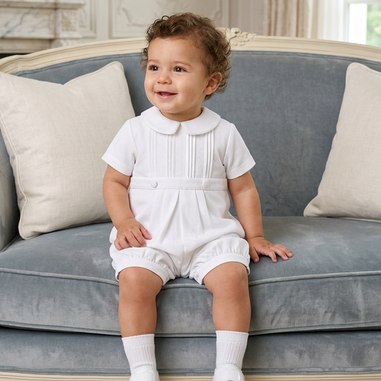 Carriage Boutique white pique baby boy romper with a Peter Pan collar, worn by a toddler sitting on a blue sofa.