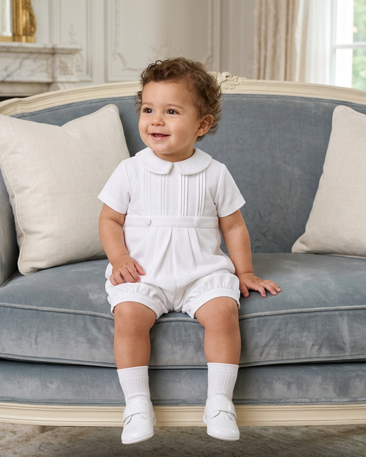 Carriage Boutique white pique baby boy romper with a Peter Pan collar, worn by a toddler sitting on a blue sofa.