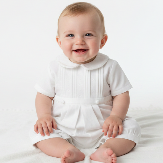 A smiling baby boy wearing a white Carriage Boutique pique romper with a peter pan collar and pleated front detail.