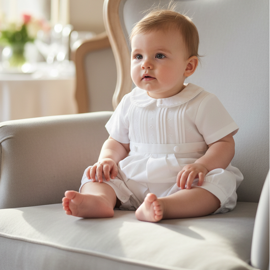 Carriage Boutique white pique baby boy romper with a Peter Pan collar, sitting on a classic upholstered armchair.