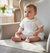 Carriage Boutique white pique baby boy romper with a Peter Pan collar, sitting on a classic upholstered armchair.