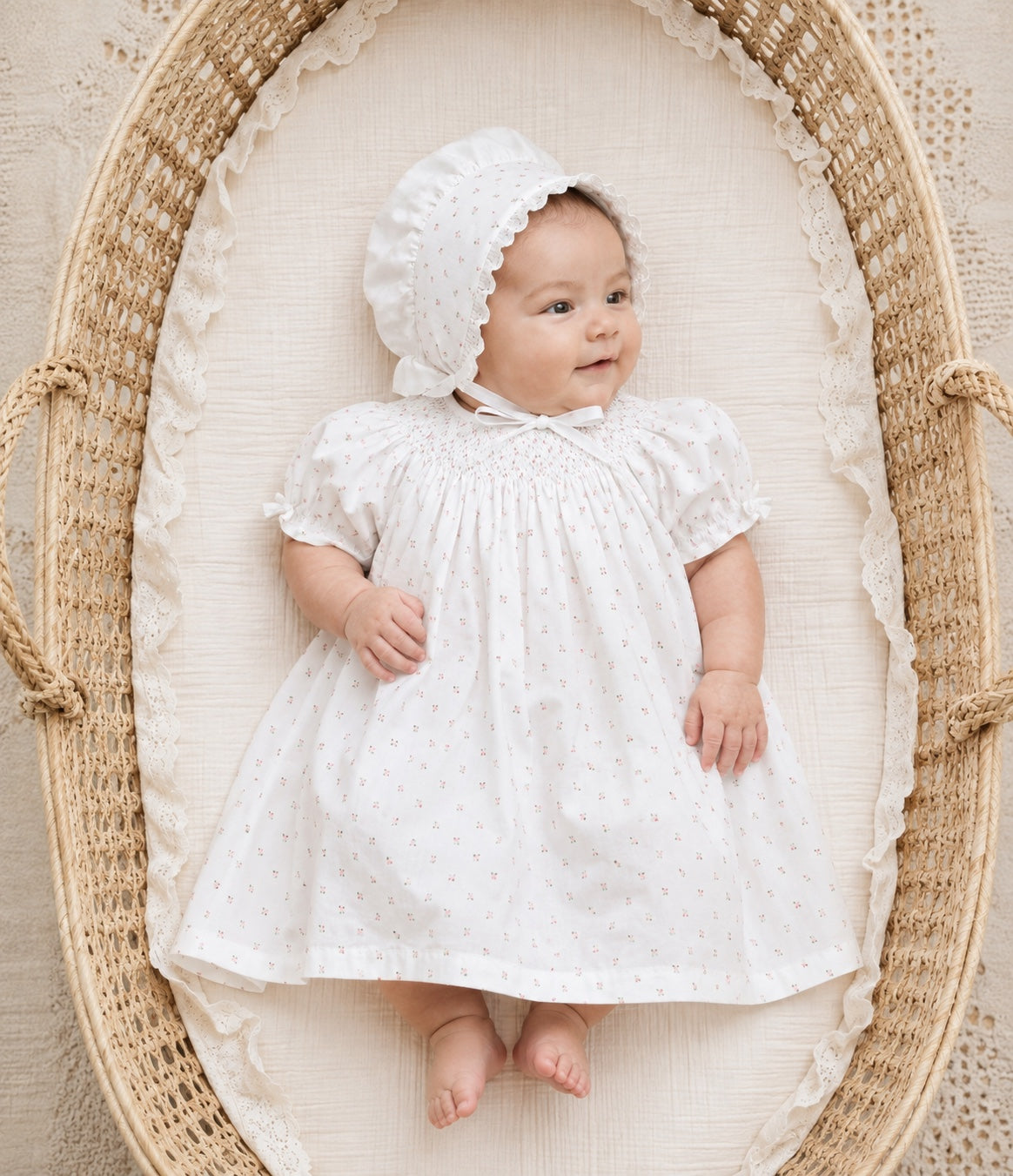 Carriage Boutique white newborn girl dress with delicate rosebud print and matching bonnet, lying in a wicker basket.