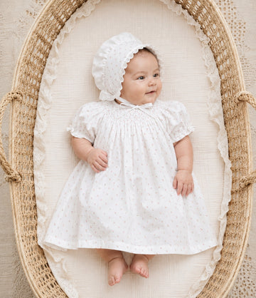Carriage Boutique white newborn girl dress with delicate rosebud print and matching bonnet, lying in a wicker basket.