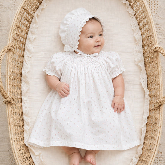 Carriage Boutique baby girl in a white rosebud print dress and matching bonnet lying in a woven wicker bassinet