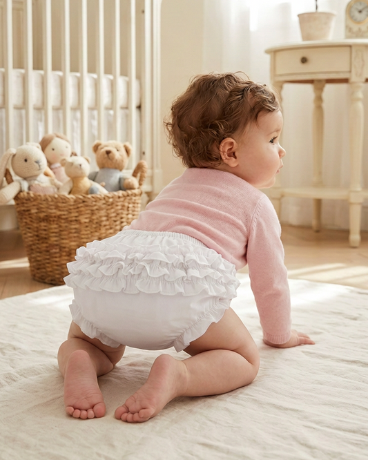 Carriage Boutique white knitted baby bloomers with ruffles on a crawling baby wearing a soft pink long-sleeve top.